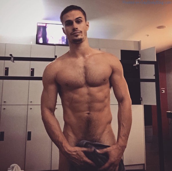 Brandy naked and posing in a locker room holding a dark towel over his manhood.