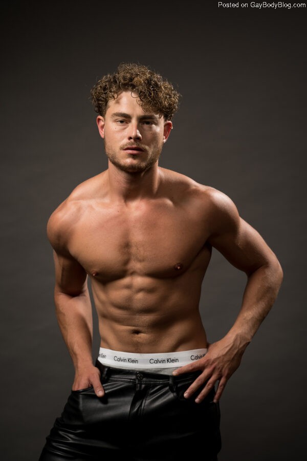 Handsome male model Haran Schechter stands shirtless against a dark background, his right hand partially in the pocket of his leather pants while his left rests on his left hip. The waistband of his Calvin Klein boxer shorts peeks out from above his leather pants.