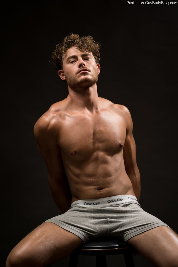 Male model Haran Schechter sits atop a stool against a black background, his chest slightly forward and his arms behind him. His athletically muscular torso is slightly obscured by shadow to display his definition. He wears only grey boxer shorts.