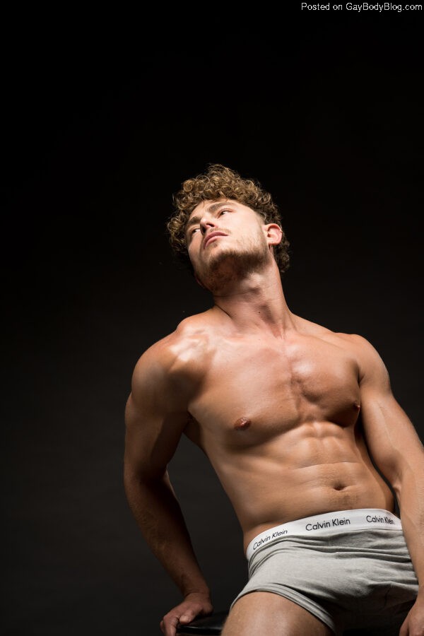 Model Haran Schechter leans back wearing only grey boxer shorts against a black background, displaying his athletically muscled body in the light