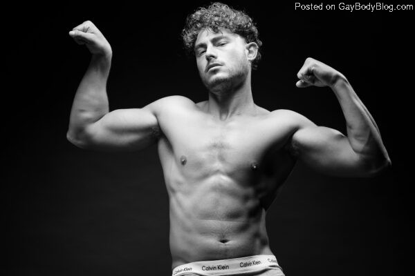 Handsome and shaggy haired German male model Haran Schechter flexes his muscles with his arms up in a black and white pose