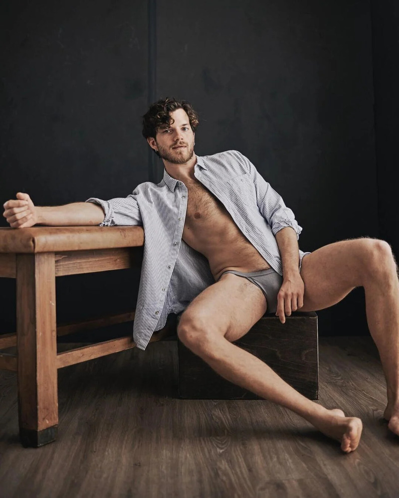 handsome model Emanuele Mariotti wearing an open blue shirt and dark grey briefs, reclining against a wooden leather-topped bench.