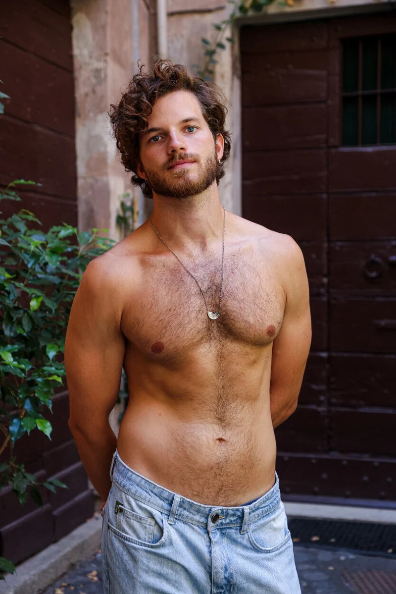 Shirtless male model Emanuele Mariotti wearing jeans and standing with his hands behind his back, showing his hairy chest.