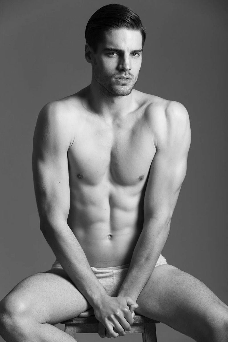 shirtless male model Kilian Isaak wearing underwear sitting on a stool and flexing his arms